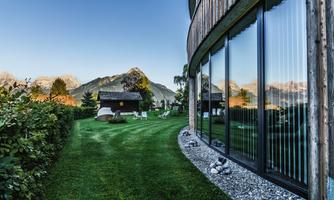 A modern building view with large windows, surrounded by green grass and mountains in the background. The garden is well-maintained and inviting.