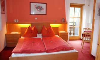 A cozy bedroom with a large bed and two red pillows. The room has orange walls and a balcony that lets in plenty of daylight.