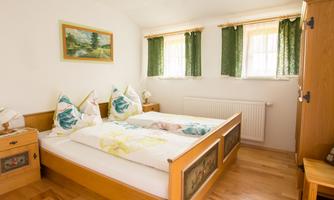 A cozy bedroom with a double bed and wooden furniture. The windows are adorned with green curtains, and a wall mural complements the décor.