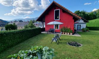 A red house with a well-kept garden and a seating area. In the background, mountains are visible under a blue sky.