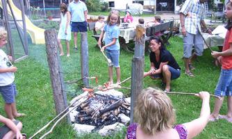 A group of children and adults gathers around a campfire outdoors. Some children roast sausages on sticks while others play in a relaxed environment.