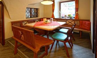 A cozy dining area with a round table and wooden benches. Large windows with curtains provide bright light and an inviting atmosphere.