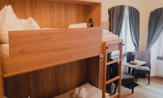 A small room with a wooden bunk bed. There is a table, chairs, and windows with curtains.