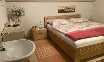 A cozy bedroom with a double bed and a light wooden dresser. Next to the bed, there is a sink on a rug.