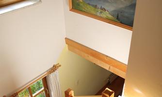 A staircase with a wooden railing leads down. A landscape painting hangs on the wall, and sunlight streams in through the window.