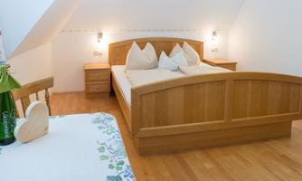 A cozy bedroom with a wooden double bed and white bed linens. A wooden chair and a table with a cloth are in the foreground.