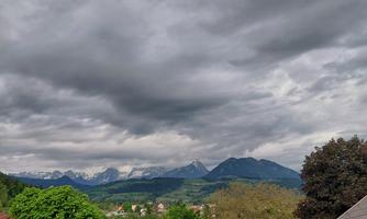 A cloudy landscape with green trees and mountains in the background. The sky is gray and suggests rain.