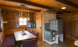 A cozy room with wooden paneling and a large dining table. In the corner, there is a tiled stove and a television is visible.