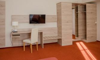A modern hotel room with a desk, a television, and a wardrobe. The floor is covered with red carpet, and the walls are brightly colored.