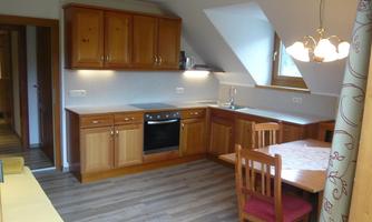 A modern kitchen with wooden cabinets and a dining area. It is bright and friendly designed, with large windows that let in daylight.