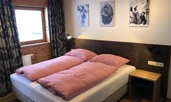 A cozy bedroom with a double bed and checked bedcovers. Some black-and-white pictures hang on the walls, and the room has large windows with curtains.