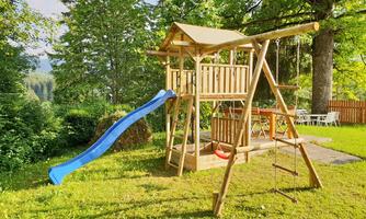 A playground with a wooden structure, a blue slide, and a swing. The green lawn and the trees in the background create an inviting atmosphere.