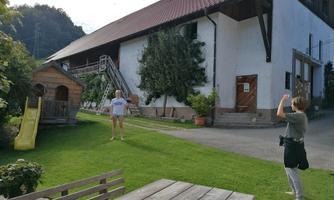 A rural scene with a large building in the background. In the foreground, two people are standing, one is taking a photo while the other is posing.