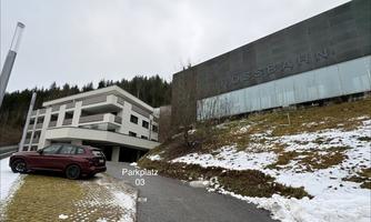 A modern building with the inscription "Hossbahn" and a parking lot in the foreground. In the background, snow-covered meadows and trees can be seen.