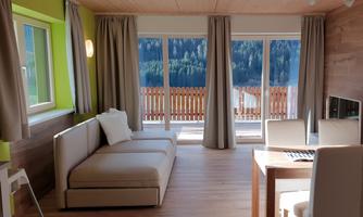 A bright and modern living room with a comfortable sofa and large windows. The view extends to a picturesque landscape with trees and a wooden terrace.