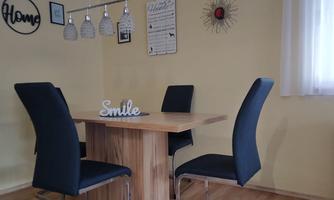 A bright dining area with a wooden strip and modern black chairs. Decorative elements hang on the wall and a friendly message is on the table.