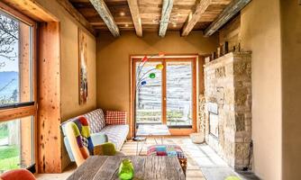 A cozy room with wooden beams and a large window that offers a view outside. The decor is modern and colorful, featuring a rustic fireplace and a wooden table.