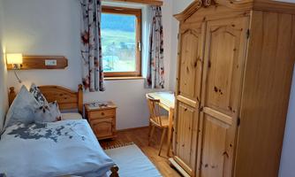 A cozy bedroom with a single bed, a wooden wardrobe, and a desk. A window overlooking the landscape brings daylight into the room.
