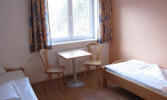 A simple bedroom with two beds and a table. The windows let in a lot of natural light.