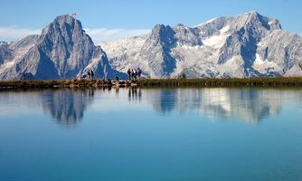 A clear lake reflects majestic mountains with snow-capped peaks. Some people are standing on the shore enjoying the view.