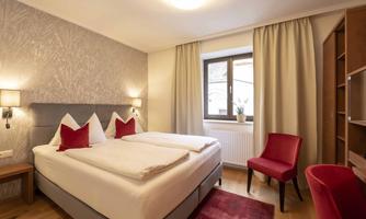 A cozy bedroom with a double bed and red cushions. The room has large windows, bright walls, and stylish furniture.