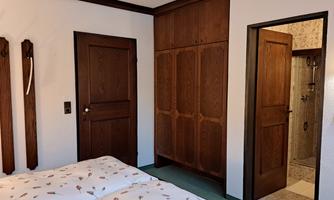 A simple bedroom with a double bed and a wooden wardrobe. In the background, a bathroom with a shower can be seen.