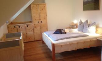 A cozy bedroom with a double bed, two pillows, and a baby crib. There is also a wooden wardrobe and warm light in the room.