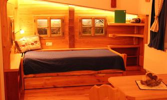 A cozy room with a wooden bed and warm light. Windows and a wooden shelf are visible on the wall.