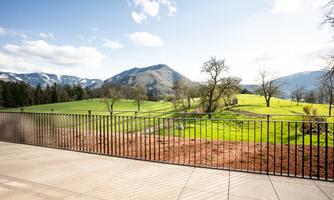 A green landscape with gentle hills and trees under a clear sky. In the foreground, an elegant railing is visible.
