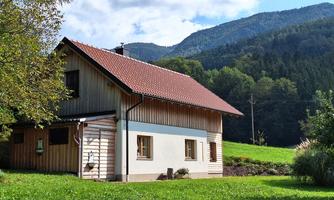 A charming wooden house in a green setting. In the background, mountains and a clear sky can be seen.