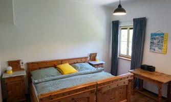 A cozy wooden bedroom with a large bed and colorful pillows. There are curtains in front of the window, and a small table with a lamp is also visible.