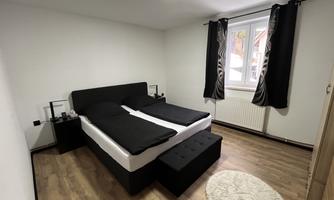 A modern bedroom with a double bed and dark blue curtains. The room has a light wooden floor and a large window.