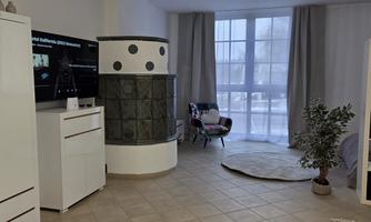A modern living space with a large window and bright curtains. In the corner stands a stove, and there is an upholstered armchair and a plant.