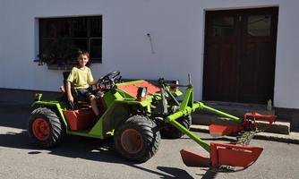 A boy is sitting on a green tractor in front of a white house. The tractor has a red shovel and a green attachment.