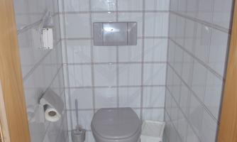 A simple bathroom with white tiles. In the middle is a toilet, next to it toilet paper and a trash can.