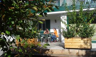 Two women sit relaxed on the terrace of a house. Surrounded by plants, they enjoy the sun and their time together.