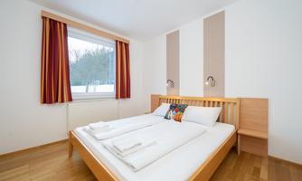 A modern bedroom with a double bed and wooden floor. Large windows let in plenty of light, and the walls are painted in bright colors.