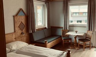 A cozy room with two beds, a bench, and a small table. Large windows provide plenty of light and an inviting atmosphere.