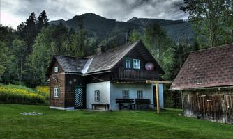 A traditional house with a dark wooden facade and balcony in a green landscape. In the background, mountains and trees can be seen.