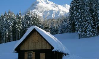 A small, traditional wooden house stands in a snowy landscape. In the background, tall mountains and dense, snow-covered trees can be seen.