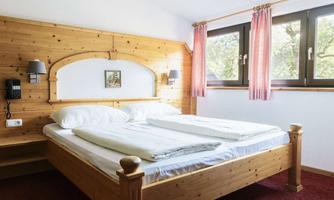 A cozy bedroom with a wooden bed and two mattresses. Large windows with red curtains and a warm atmosphere are present.