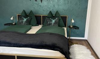 A modern bedroom with a green wall and a mountain pattern. The bed is dressed with dark bed linen and decorative cushions in a similar shade.