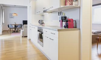 A modern kitchen with white cabinets, a stove, and a coffee machine. In the background, a cozy dining area is visible.
