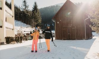 Two skiers are walking through the snow, surrounded by a picturesque winter forest. The sun is shining and a wooden house is visible in the background.