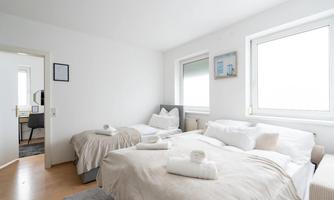 A modern bedroom with two beds and bright, neutral colors. Large windows provide plenty of daylight and a pleasant atmosphere.