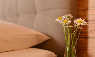 A cozy bedroom with a gray headboard and a pillow. A glass with daisies is on the nightstand.