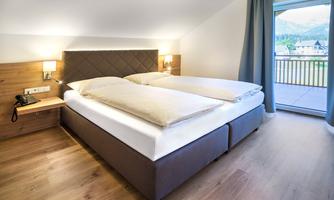 A modern bedroom with a large double bed and wooden elements. Large windows offer a view of the mountains and the outdoor area.