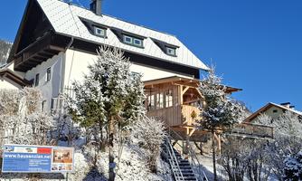 A cozy house in a winter landscape with snow and a clear blue sky. In the foreground, there are green trees and a staircase leading to the porch.
