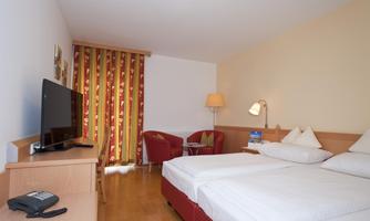 A bright hotel room with two single beds, a television, and a table. Red curtains and a cozy armchair create a pleasant atmosphere.