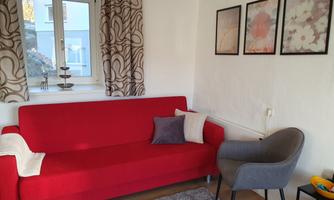 A cozy living room with a red sofa and a gray armchair. Decorative pictures are hanging on the wall, and there are curtains that filter the light.